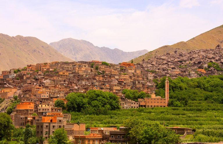 Village in the Ourika Valley, Atlas Mountains of Morocco, featuring stone houses, lush greenery, a tall minaret, and rugged mountain peaks in the background.