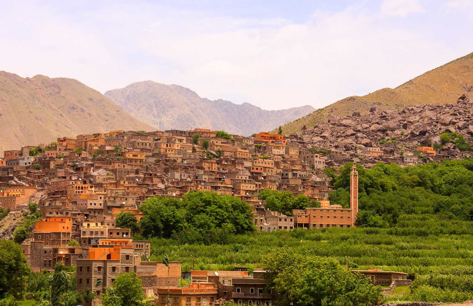 Village in the Ourika Valley, Atlas Mountains of Morocco, featuring stone houses, lush greenery, a tall minaret, and rugged mountain peaks in the background.